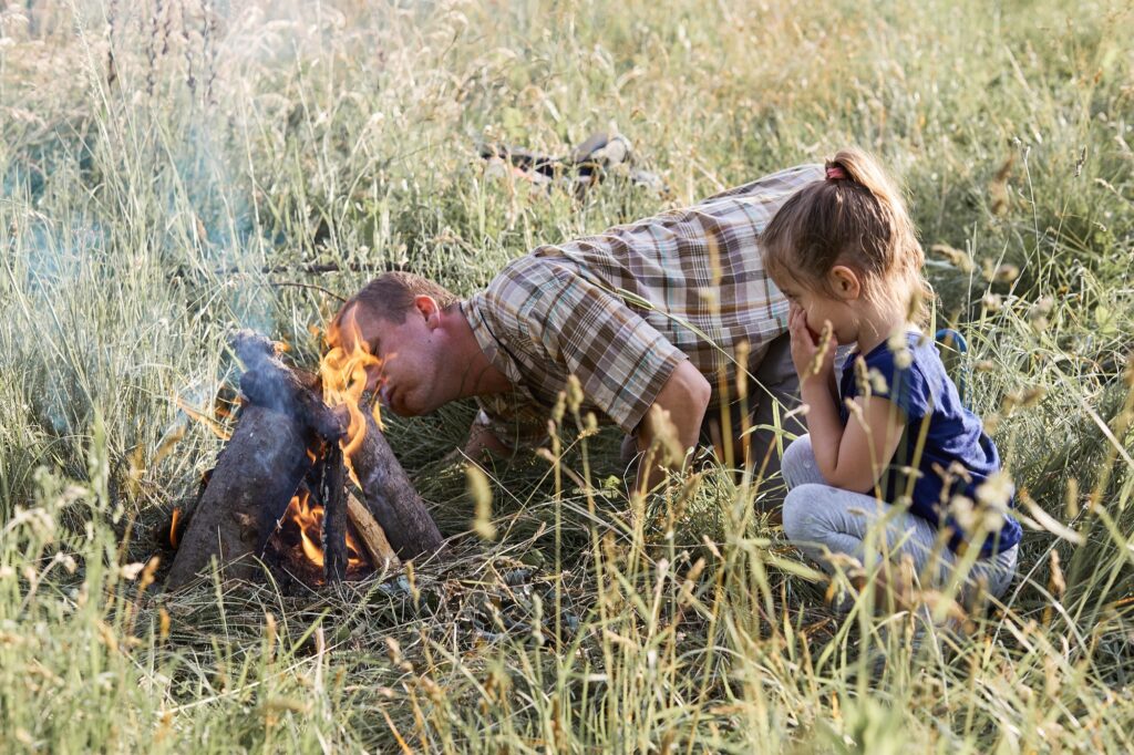 Man starting a campfire, blowing on a fire. Little girl sitting in a grass beside a campfire